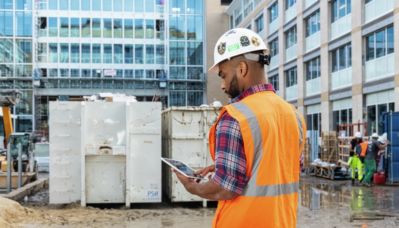 Een man op een werf met een helm op en een veiligheidsvest aan raadpleegt een tablet met de Fieldwire-toepassing.